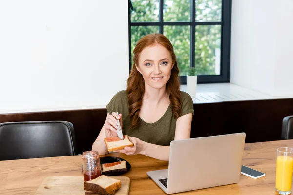 Femme souriant à la caméra tout en tenant un toast avec de la confiture près du jus d'orange et des appareils numériques sur la table — Photo de stock