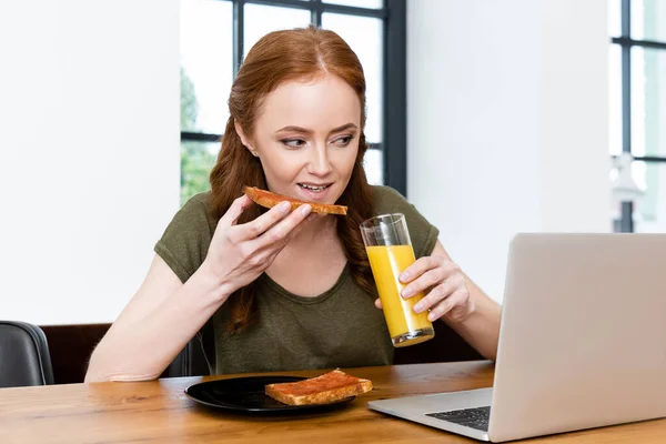 Mulher comendo torrada e segurando vidro de suco de laranja perto de laptop na mesa de madeira — Fotografia de Stock