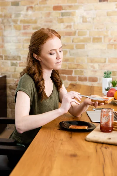Selective focus of woman adding jam on toast near digital devices and fruits on table — Stock Photo
