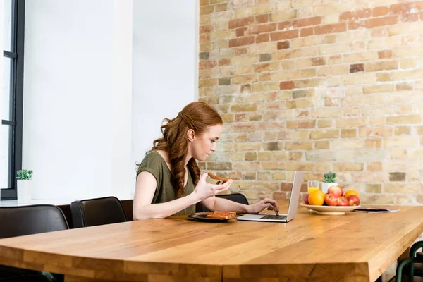 Selective focus of woman holding toast and using laptop near fruits on table — Stock Photo