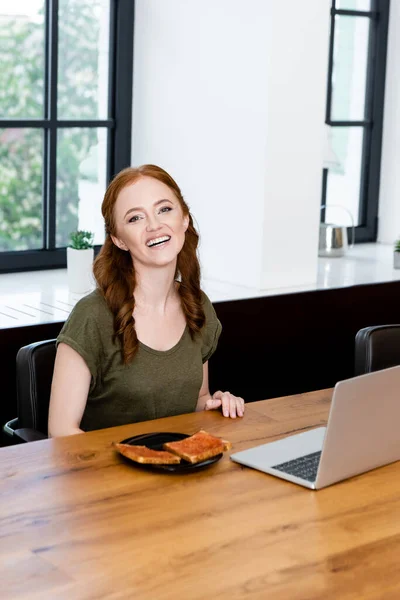 Atractiva mujer sonriendo a la cámara cerca de brindis con mermelada y portátil en mesa de madera - foto de stock
