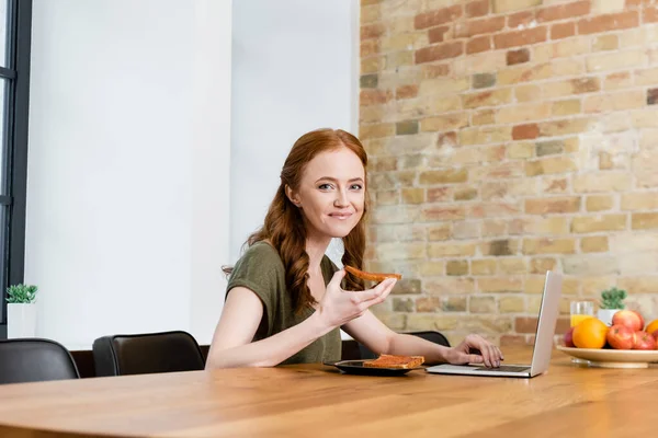 Selective focus of beautiful woman smiling at camera while holding toast and using laptop at table — Stock Photo