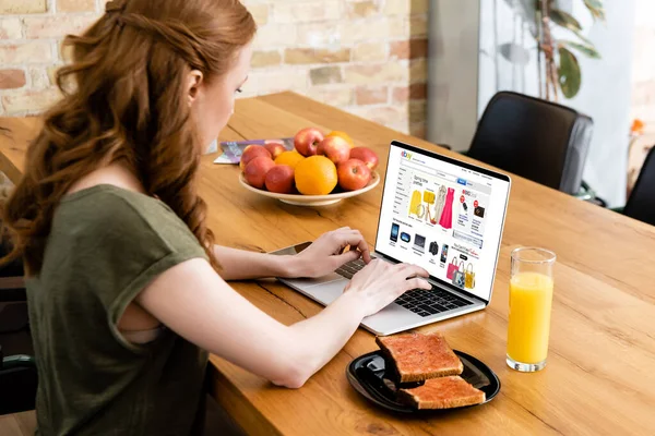KYIV, UCRANIA - 8 de mayo de 2020: Vista lateral de la mujer que usa el ordenador portátil con el sitio web de ebay cerca del vaso de jugo de naranja y tostadas con mermelada en la mesa - foto de stock