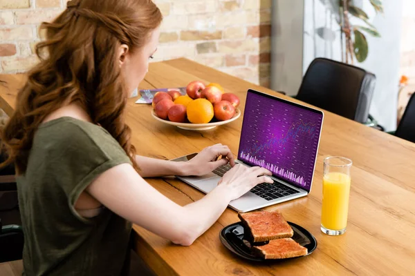 Vista lateral de la mujer usando el ordenador portátil con gráficos cerca de tostadas y jugo de naranja en la mesa - foto de stock