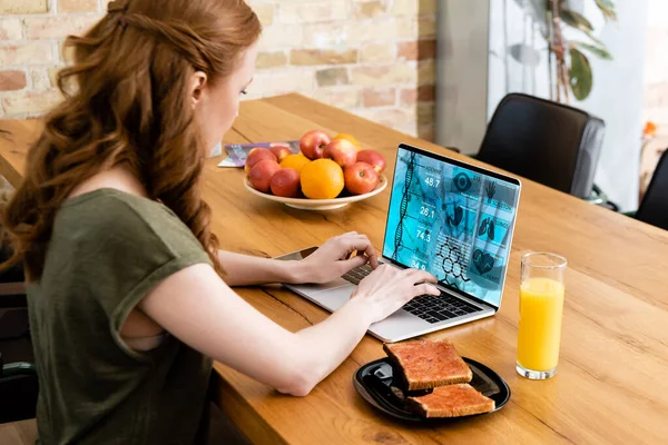 Vista laterale della donna che utilizza il computer portatile con e sito web salute vicino a vetro di succo d'arancia e toast sul tavolo di legno — Foto stock