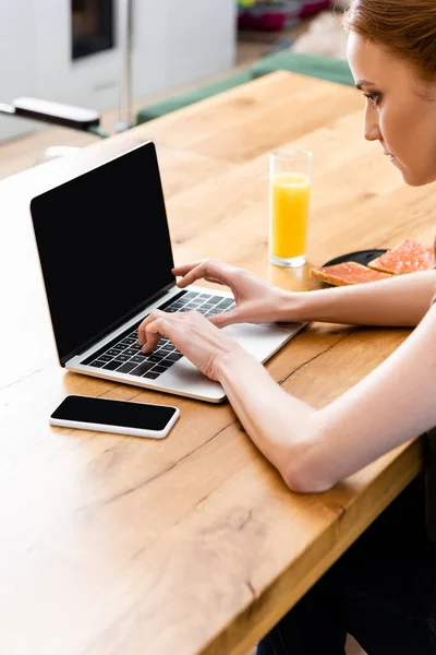 Freelancer usando laptop perto de smartphone, torradas e vidro de suco de laranja na mesa — Fotografia de Stock