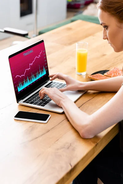 Frau benutzt Laptop mit Handelswebseite in der Nähe von Smartphone, Toast und Glas Orangensaft auf Holztisch — Stockfoto