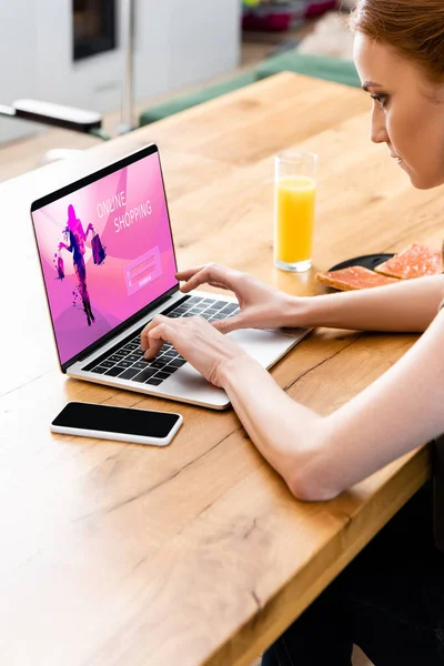 Mulher usando laptop com site de compras on-line perto de smartphone e café da manhã na mesa — Fotografia de Stock