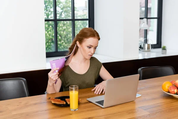 Teletrabajador agitando revista mientras utiliza el ordenador portátil cerca del desayuno en la mesa de madera - foto de stock