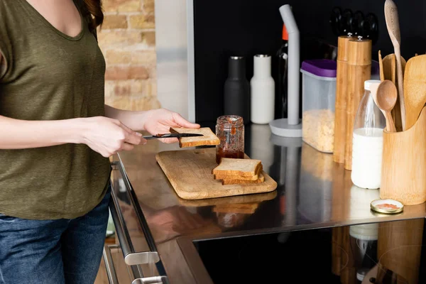 Cropped view of woman pouring jam on toast on kitchen worktop — Stock Photo