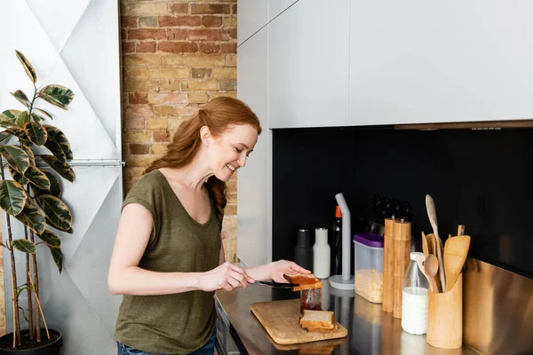 Lächelnde Frau gießt Marmelade auf Toast in der Nähe der Küchenarbeitsplatte — Stockfoto