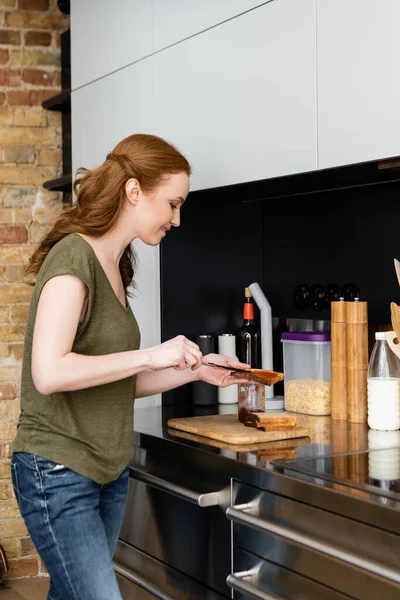 Vue latérale de la femme souriante versant de la confiture sucrée sur du pain grillé près du plan de travail de la cuisine — Photo de stock