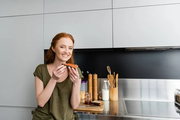 Bella donna sorridente alla macchina fotografica mentre tiene il pane tostato con marmellata in cucina — Foto stock