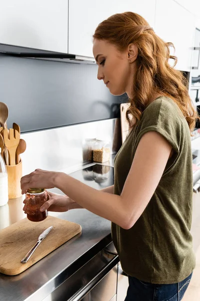 Vista lateral de mujer atractiva sosteniendo tarro con mermelada en la cocina - foto de stock