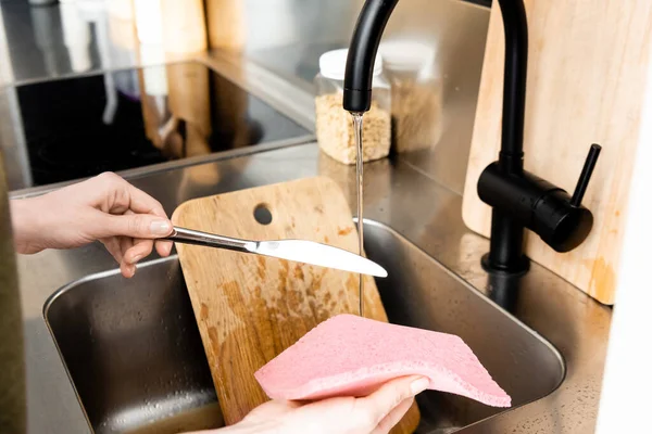 Обрезанный вид женщины, держащей тряпку и нож возле кухонного крана и раковины — стоковое фото