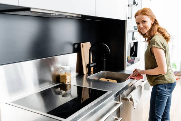 Vue latérale de la femme souriant à la caméra tout en nettoyant le plan de travail de la cuisine avec chiffon — Photo de stock