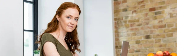 Panoramic crop of woman looking at camera near laptop at home — Stock Photo