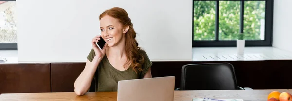 Панорамний урожай усміхненої жінки, що говорить на смартфоні біля ноутбука на столі вдома — стокове фото