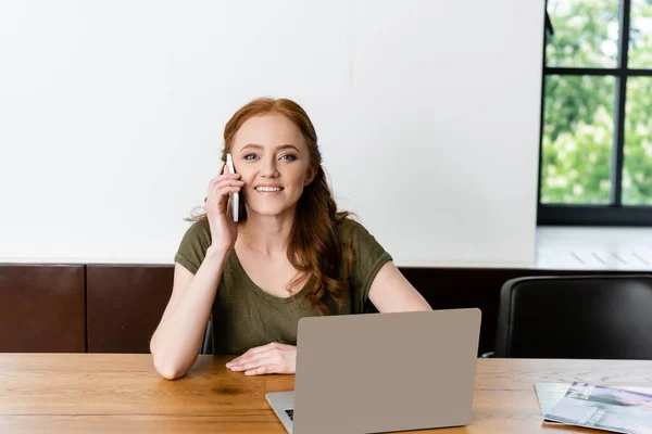 Freelancer sonriente mirando a la cámara mientras habla en el teléfono inteligente cerca de la computadora portátil en la mesa - foto de stock