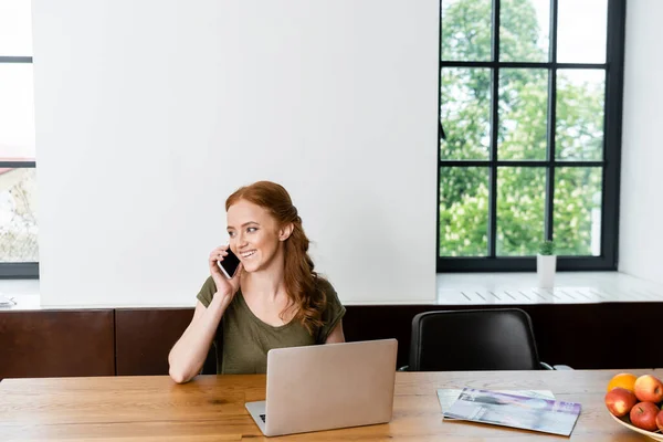 Mujer atractiva sonriendo mientras habla en el teléfono inteligente cerca de la computadora portátil, revistas y frutas en la mesa - foto de stock