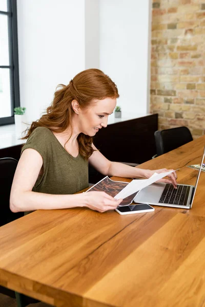 Foco seletivo de freelancer sorridente trabalhando com papéis e laptop na mesa — Fotografia de Stock