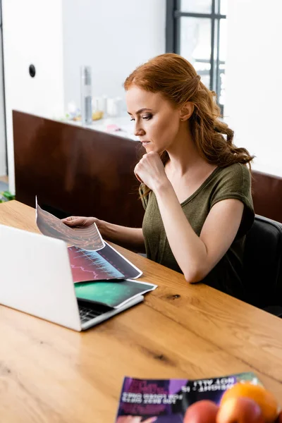 Selective focus of freelancer holding paper with charts near laptop, magazine and fruits on table — Stock Photo