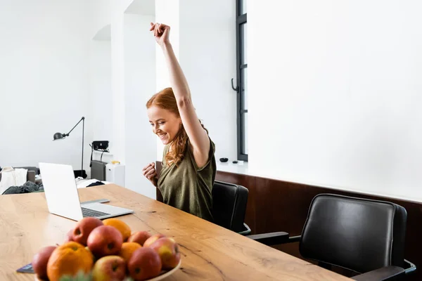 Focus selettivo di donna allegra mostrando sì segno mentre seduto a tavola vicino al computer portatile — Foto stock