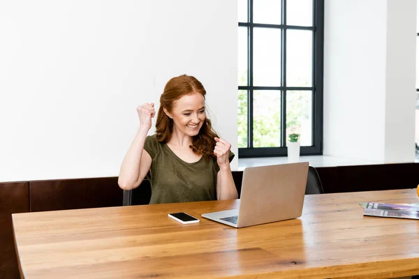 Smiling woman showing yeah gesture near laptop and smartphone on table at home — Stock Photo