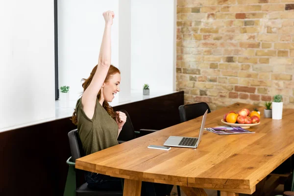 Вид сбоку жизнерадостной женщины, показывающей жест рядом с ноутбуком и смартфоном на столе дома — стоковое фото