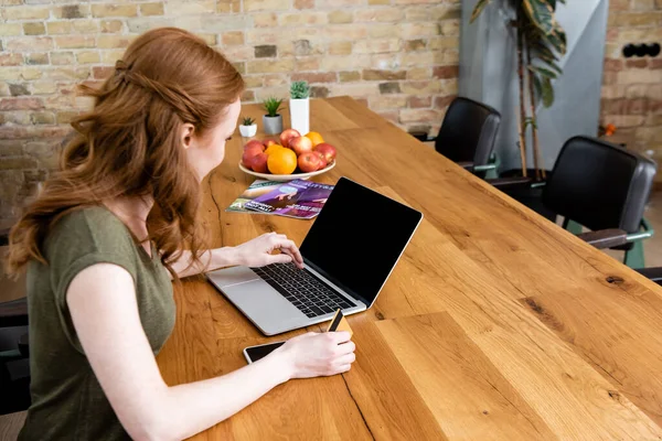 Vista lateral del freelancer usando el ordenador portátil mientras sostiene la tarjeta de crédito en la mesa - foto de stock