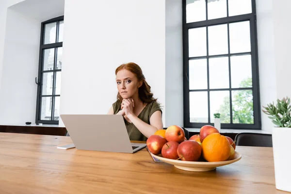 Selective focus of attractive freelancer looking at camera near digital devices, fruits and plant on table — Stock Photo