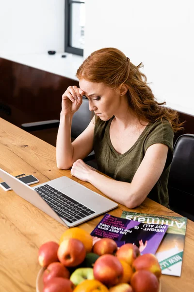 Selective focus of pensive teleworker looking at laptop near magazines and fruits on table — Stock Photo