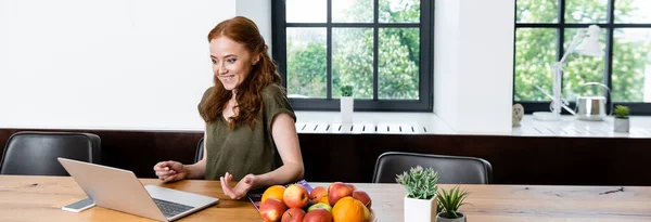 Vista panorámica de la mujer alegre teniendo video chat en el ordenador portátil cerca de frutas y plantas en la mesa - foto de stock