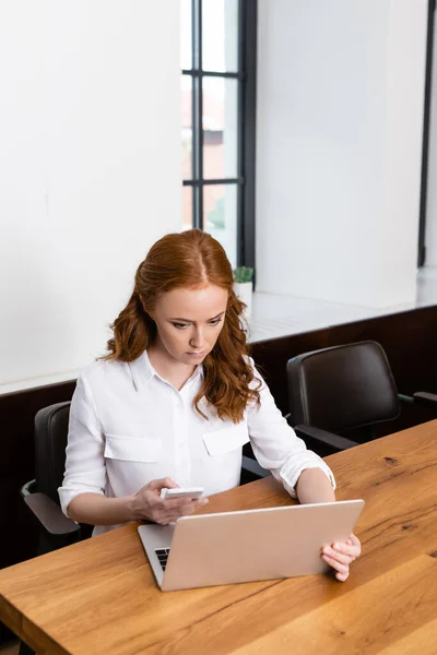 Attraktive Frau mit Smartphone und Laptop am Holztisch — Stockfoto