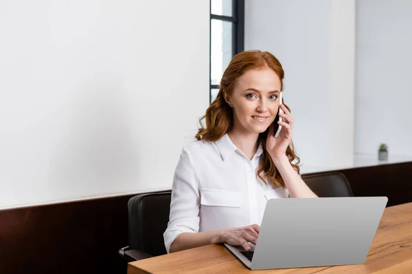 Freelance sorridente che parla su smartphone vicino a computer portatile sul tavolo — Foto stock