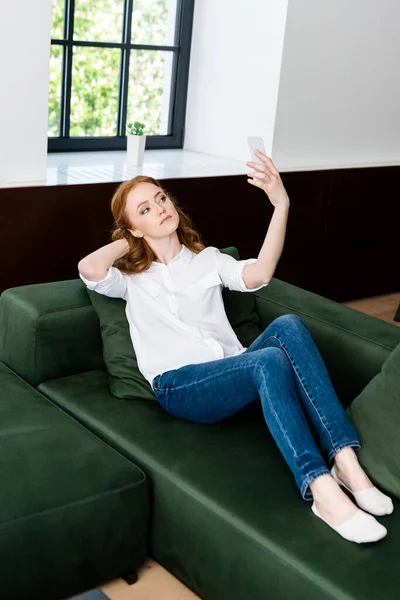 Mujer atractiva tomando selfie con teléfono inteligente en el sofá en casa - foto de stock
