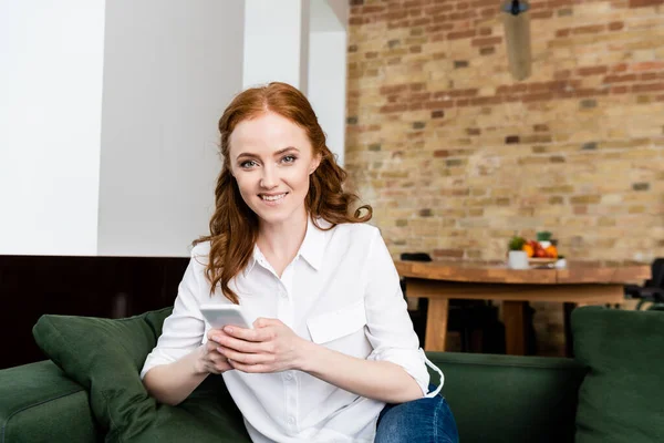 Schöne Frau lächelt in die Kamera, während sie ihr Smartphone zu Hause auf der Couch benutzt — Stockfoto