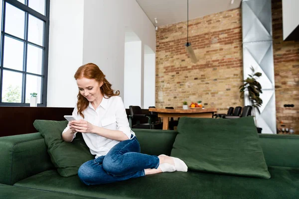 Усміхнена руда волохата жінка використовує смартфон, сидячи на дивані вдома — стокове фото