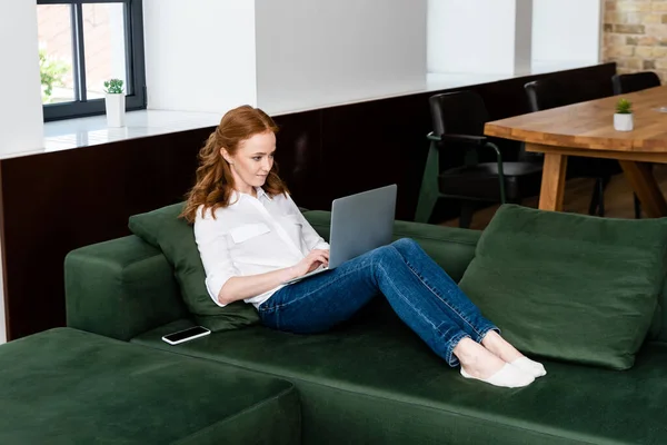 Attractive teleworker using laptop near smartphone on couch in living room — Stock Photo