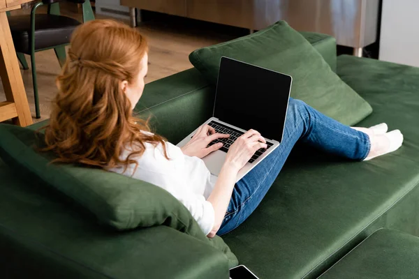 Mise au point sélective de pigiste aux cheveux roux à l'aide d'un ordinateur portable près du smartphone sur le canapé — Photo de stock