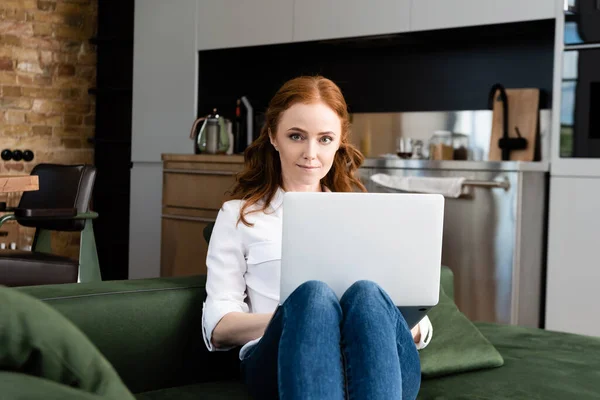 Selective focus of attractive freelancer looking at camera while using laptop on sofa in living room — Stock Photo