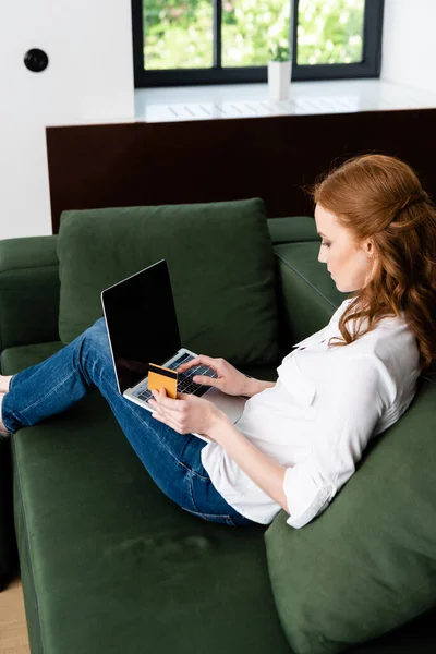 Vista lateral de la mujer usando el ordenador portátil y la tarjeta de crédito en el sofá en casa - foto de stock