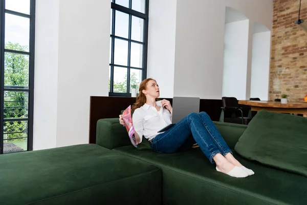 Attractive woman holding magazine and laptop while suffering from heat on sofa in living room — Stock Photo