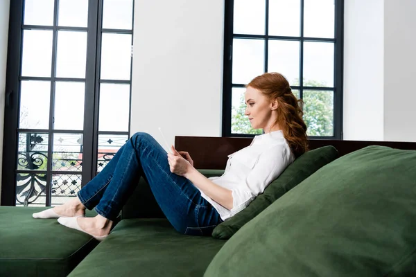 Вид збоку красивої червоної волохатої жінки з використанням цифрового планшета на дивані — стокове фото