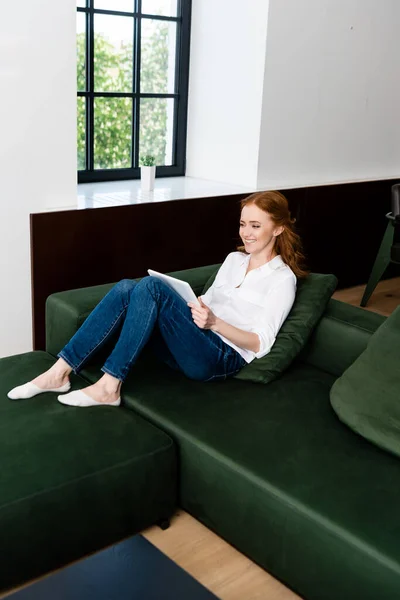 Mujer sonriente usando tableta digital mientras está sentada en el sofá en la sala de estar - foto de stock