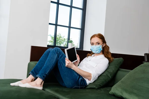 Femme en masque médical montrant tablette numérique avec écran blanc sur le canapé — Photo de stock