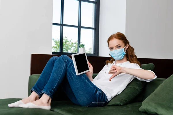 Woman in medical mask showing thumb down and holding digital tablet on couch — Stock Photo