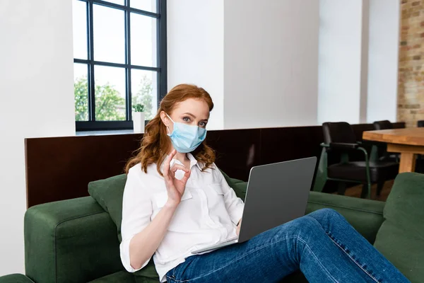 Frau mit medizinischer Maske zeigt Okay-Geste, während sie zu Hause Laptop benutzt — Stockfoto