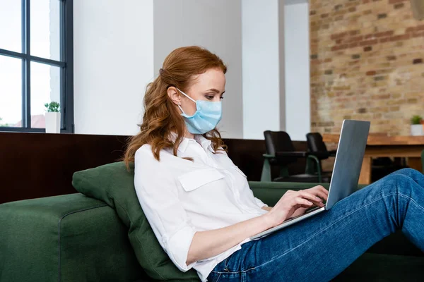 Vista lateral del teletrabajador en máscara médica utilizando el ordenador portátil en el sofá - foto de stock
