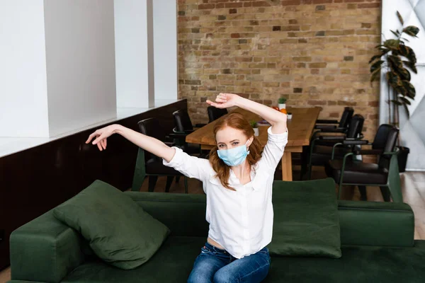 Femme en masque médical avec les mains levées regardant la caméra sur le canapé — Photo de stock